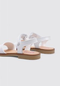 Sandalo bianco con design a punta aperta, cinturino in tessuto, chiusura regolabile in velcro, suola piatta marrone e dettaglio stampato nella soletta.