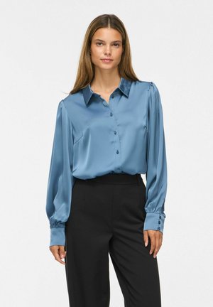 Femme aux longs cheveux lisses portant un chemisier bleu en satin boutonné et un pantalon noir taille haute, debout devant un fond uni.
