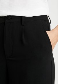 Zwarte op maat gemaakte pantalon van een gladde stof. Voorzien van een knoopsluiting, zijzakken en een strakke, getailleerde pasvorm.