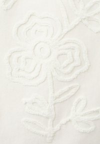 Broderie florale blanche en relief avec des pétales et des feuilles texturés sur un fond de tissu clair, créant un motif décoratif délicat.