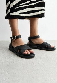 Sandalias de cuero negras con correas cruzadas, superficie texturizada, correa de tobillo con hebilla y suela de goma plana. Combinadas con una falda de patrón en blanco y negro.