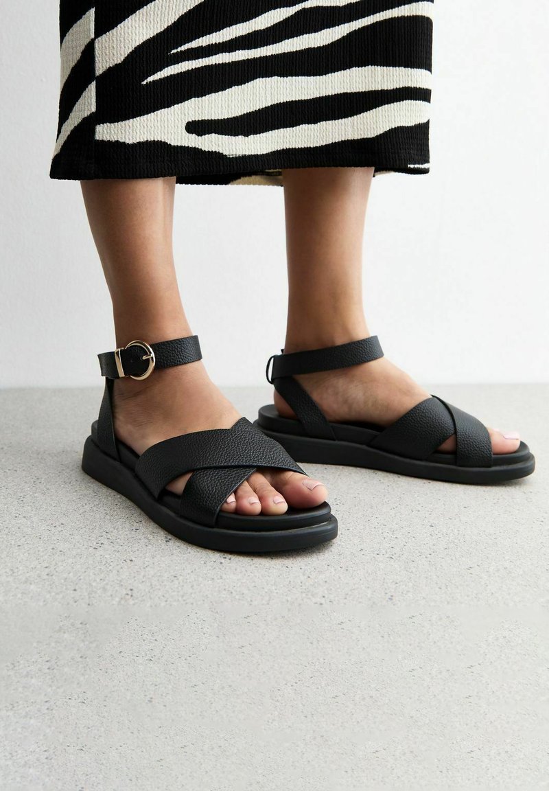 Sandalias de cuero negras con correas cruzadas, superficie texturizada, correa de tobillo con hebilla y suela de goma plana. Combinadas con una falda de patrón en blanco y negro.