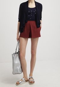 Marinefarbener leichter Cardigan über einem marinefarbenen Tanktop, kombiniert mit rostfarbenen, gefalteten Shorts. Silberne Sandalen und eine strukturierte silberne Handtasche.