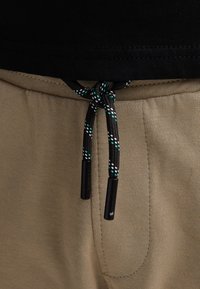 Beige shorts med en sort linning og en flettet snøre i sort med grønne og hvide accenter, fastgjort med plastiktipper.