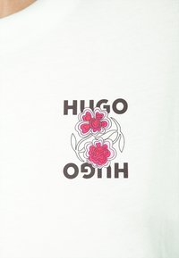 Bílá bavlněná tričko s grafickým designem dvou růžových květin a textem "HUGO" v tučných černých písmenech, zobrazený vertikálně.