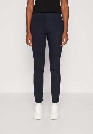 Femme portant un pantalon slim bleu marine foncé, un haut sans manches noir et des baskets blanches, debout devant un fond clair uni.