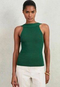 Grünes, geripptes, ärmelloses Oberteil mit hohem Ausschnitt, das ein enganliegendes Design und Aussparungen an den Schultern aufweist, kombiniert mit cremefarbenen Hosen.