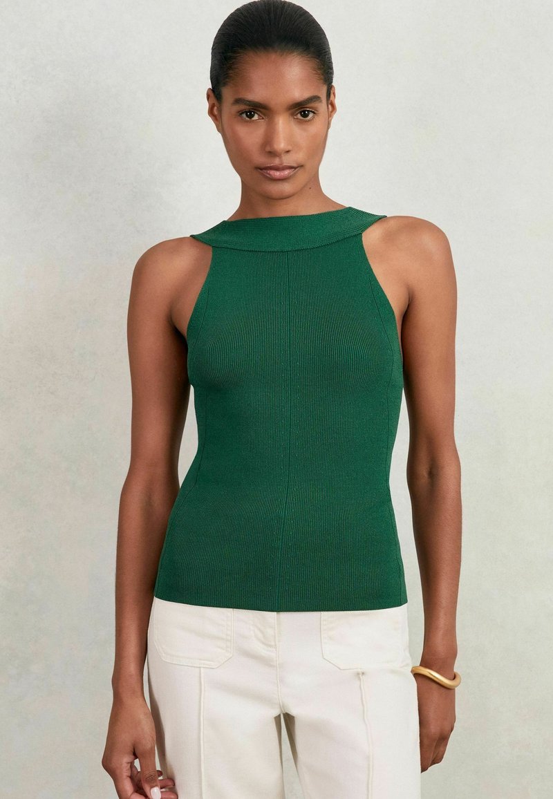 Grünes, geripptes, ärmelloses Oberteil mit hohem Ausschnitt, das ein enganliegendes Design und Aussparungen an den Schultern aufweist, kombiniert mit cremefarbenen Hosen.