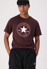 T-shirt en coton marron avec une étoile blanche et un logo rouge "Converse All Star". Manches courtes, col rond, coupe décontractée. Porté avec un short noir.