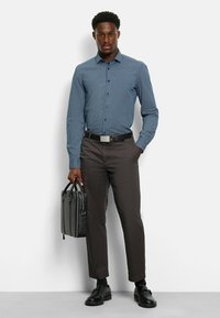 Hemd in Marineblau mit einem kleinen weißen geometrischen Muster, kombiniert mit dunkelgrauen Hosen und schwarzen Schuhen. Er trägt eine schwarze Aktentasche.