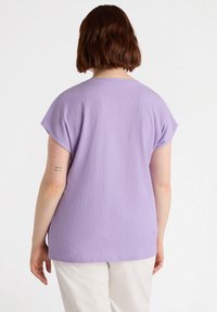 Laventelin lyhythihainen paita, jossa on tekstuuripinta, pyöreä kaula-aukko ja rento leikkaus. Yhdistetty vaaleisiin housuihin, esittäen yksinkertaista muotoilua.