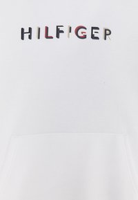 Felpa bianca con cappuccio, dotata di una tasca frontale e ricamo "HILFIGER" in vari colori: nero, rosso, beige e blu navy. Materiale morbido in misto cotone.