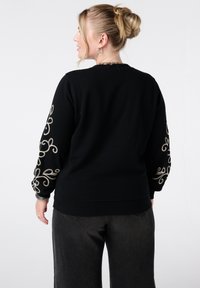 Zwarte pullovertrui met ingewikkelde gouden borduursels op de mouwen, geribbelde onderkant en manchetten. Gecombineerd met donkere, wijde broek.