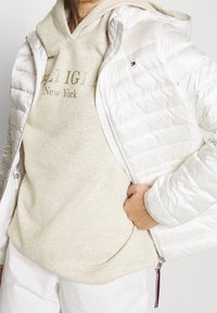 Chaqueta blanca acolchada con capucha y cierre de cremallera, llevada sobre un sudadera beige con el texto "Tommy Hilfiger New York", con un fondo neutro.