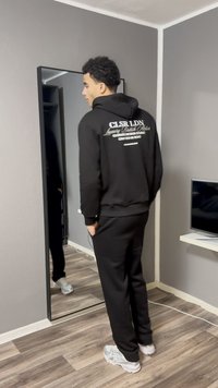 Μαύρο φούτερ με κουκούλα και λευκό κείμενο στην πλάτη, συνδυασμένο με μαύρο παντελόνι φόρμας. Στέκεται μπροστά σε καθρέφτη, με λεπτή σχεδίαση.