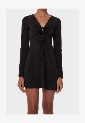 Femme portant une mini-robe noire à manches longues avec un devant froncé et un nœud noué au niveau de l'encolure.