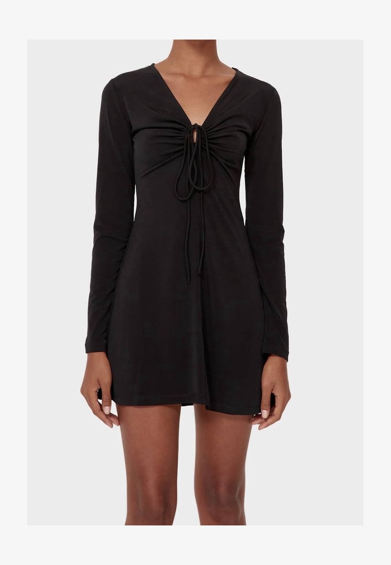 Femme portant une mini-robe noire à manches longues avec un devant froncé et un nœud noué au niveau de l'encolure.