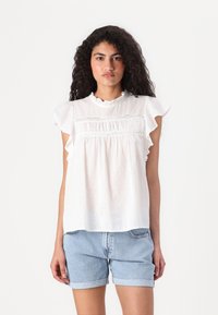 Blouse blanche avec des manches à volants, tissu texturé avec des pois, détails en dentelle au niveau de la poitrine, associée à un short en jean.