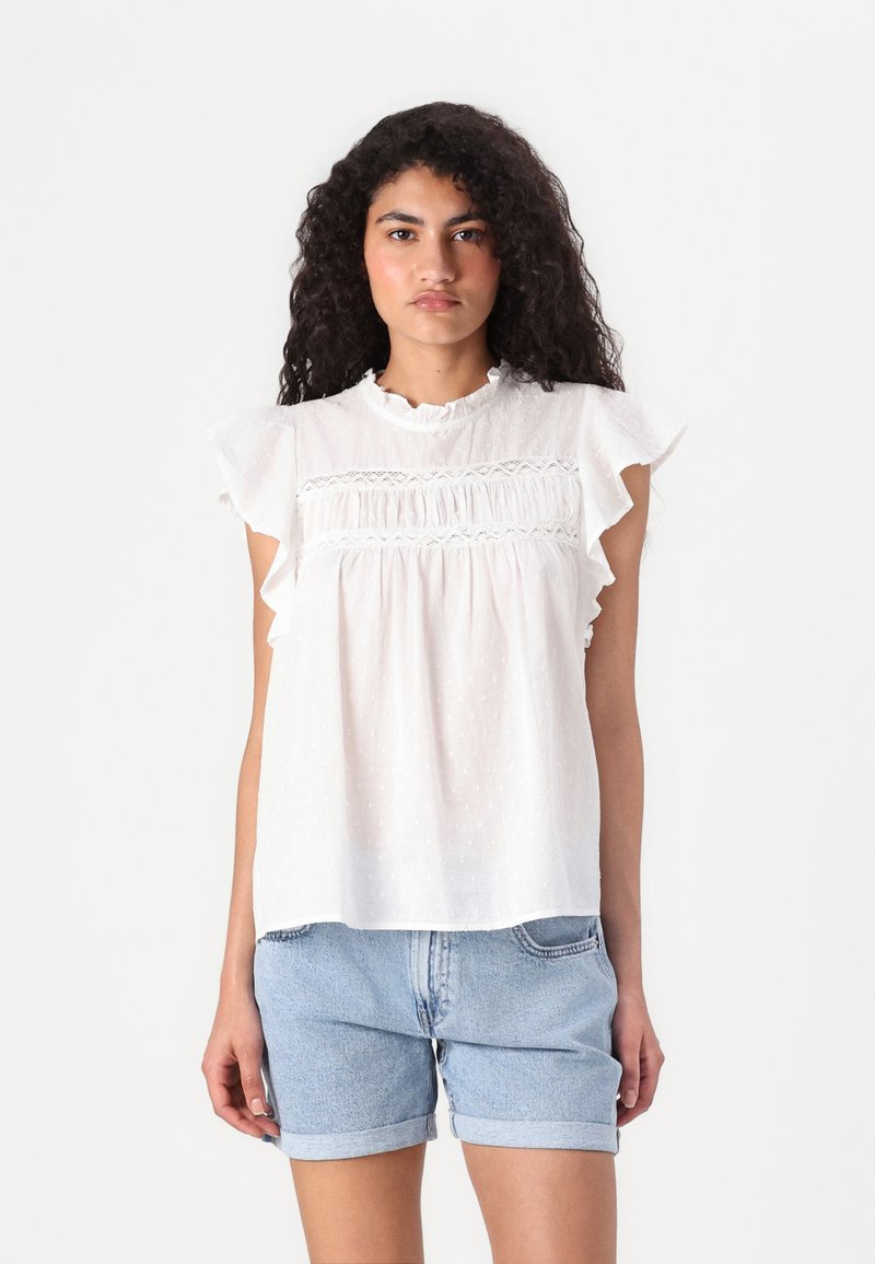 Blouse blanche avec des manches à volants, tissu texturé avec des pois, détails en dentelle au niveau de la poitrine, associée à un short en jean.