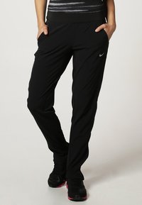 Black athletic pants with a tapered leg, elastic waistband, side pockets, and a reflective Nike logo on the left side. Smooth, lightweight fabric.