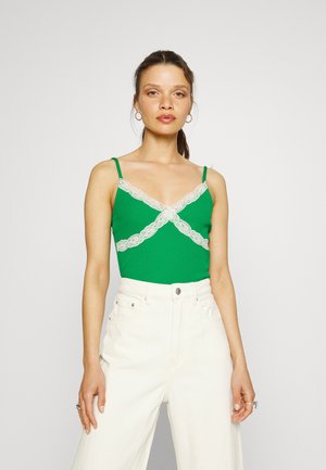 Top verde com rendas na gola, apresenta alças finas e um corpo ajustado. Usado com calças brancas de cintura alta.