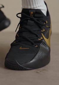 Black athletic shoe with gold accents, textured material, and a prominent Nike swoosh. Features black laces and a patterned sole.
