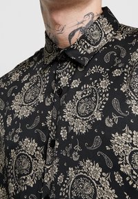 Černá košile s krátkým rukávem zdobená paisley a květinovým vzorem v béžové barvě. Vyrobena z hladké tkaniny s límečkem a černými knoflíky.