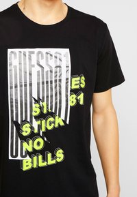 Sort bomuld t-shirt med et grafisktryk i hvid og neon grøn tekst, der viser "GUESS81" og "STICK NO BILLS" med en revnet effekt.