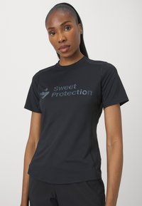 Svart kortermet treningsskjorte laget av pustende materiale, med "Sweet Protection"-logoen i en litt blank trykk på forsiden.