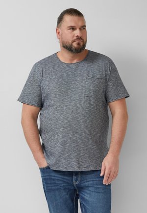 Mann mit Bart und zurückgebundenen Haaren, trägt ein graues T-Shirt mit kurzen Ärmeln und blaue Jeans, steht vor einem schlichten hellen Hintergrund.