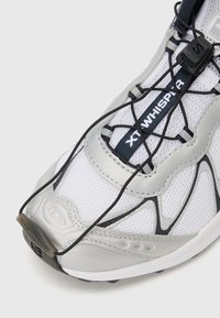 Bijela i srebrna sportska cipela s mrežastim gornjim dijelom, elastičnim vezicama i teksturiranim potplatom s zaobljenim vrhom i detaljem logotipa.