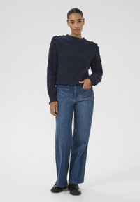 Navy knitted sweater with ribbed texture and ruffled side details, paired with wide-leg blue denim jeans and black shoes.