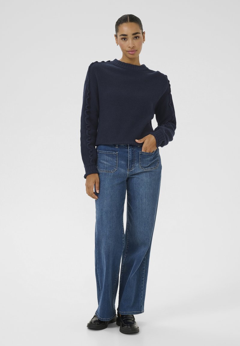 Navy knitted sweater with ribbed texture and ruffled side details, paired with wide-leg blue denim jeans and black shoes.