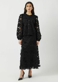 Svart brodert to-delt antrekk med en gjennomsiktig, blomsterdekorert langermet topp og matchende skjørt, kombinert med svarte høye hæler.