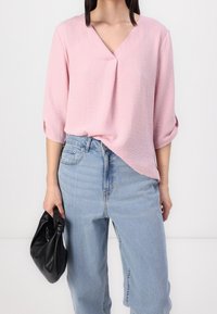 Ljusrosa, strukturerad blus med V-urringning och trekvartslånga ärmar, kombinerad med högmidjade denimjeans och en svart kuvertväska.