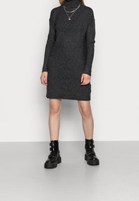 Robe tricotée gris foncé avec des manches longues et un col roulé. Portée avec des bottines noires chunky avec des accents de sangle. Design simple et ajusté.