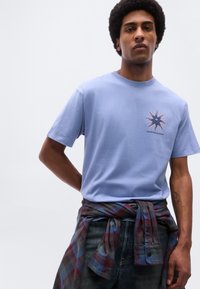 Νεαρός με αφροχτένισμα, φοράει ανοιχτό γαλάζιο μπλουζάκι με λογότυπο αστέρι, πουκάμισο καρό δεμένο γύρω από τη μέση και σκούρο τζιν.