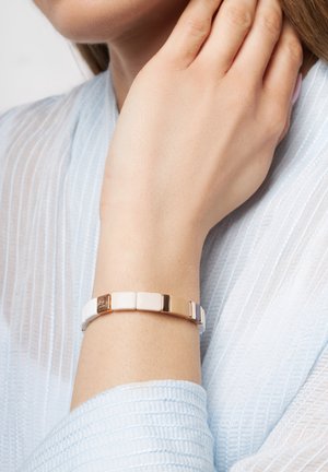 Weißes und goldenes Armband mit quadratischen Gliedern, abwechselnd glänzend weiß und metallischen Akzenten, getragen an einer Hand vor einem hellblauen Hintergrund.
