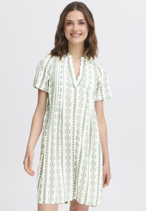 Jeune femme aux cheveux bruns mi-longs, portant une robe blanche longueur genoux avec des motifs verts verticaux, souriante, debout devant un fond uni.