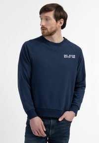 Marineblå sweatshirt med rund hals, raglanærmer og tekst "Drei Master" på brystet. Fremstillet af blødt stof med ribbede manchetter.