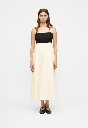 Žena v čierno-bielych maxi šatách bez rukávov s tenkými ramienkami stojí pred jednoduchým svetlošedým pozadím.