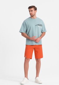 T-shirt en coton bleu clair avec un design graphique sur la poitrine, associé à un short orange. Le mannequin porte des baskets blanches et se tient dans une pose neutre.