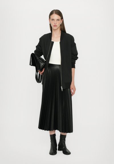Giubbotto bomber nero con zip, top bianco e lungo gonna plissettata nera; modella che tiene una borsa a tracolla in pelle nera; stivali neri al ginocchio.