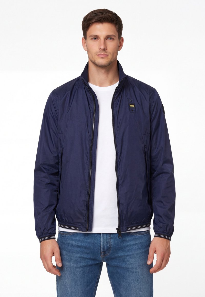 Giovane uomo che indossa una giacca blu navy con cerniera sopra una t-shirt bianca e jeans blu, in piedi davanti a uno sfondo bianco semplice.