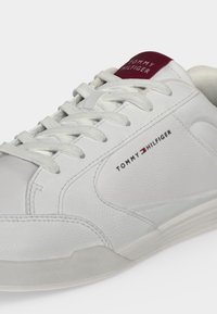 Λευκό δερμάτινο αθλητικό παπούτσι Tommy Hilfiger με λευκά κορδόνια, λογότυπο στο πλάι και βυσσινί ετικέτα γλώσσας με το εμπορικό σήμα.