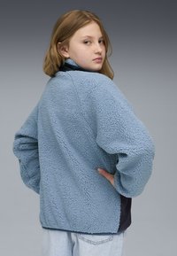 Chaqueta de forro polar en azul claro, con una superficie texturizada, corte holgado y acentos en azul marino oscuro en el dobladillo y el cuello.