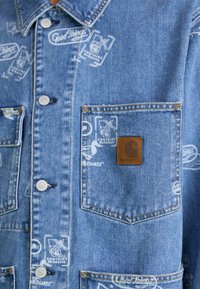 Light blue denim jacket featuring white graphics and a brown leather logo patch on the chest pocket. Two front pockets with button closures.
