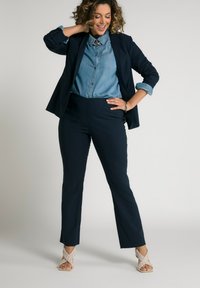 Marinblå blazer över en ljus denimskjorta, kombinerad med avsmalnande marinblå byxor och beige vävda sandaler. Slät textil och skräddarsydd passform.