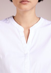 Vit bomullsskjorta med rund halsringning, subtil lyster och en knappslå. Slät yta, ärmar i medellängd och minimalistisk design.