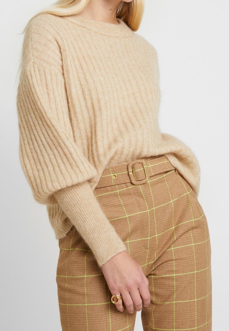 Maglione beige a coste con maniche ampie, abbinato a pantaloni a quadri marroni con accenti gialli e una cintura. Texture liscia.